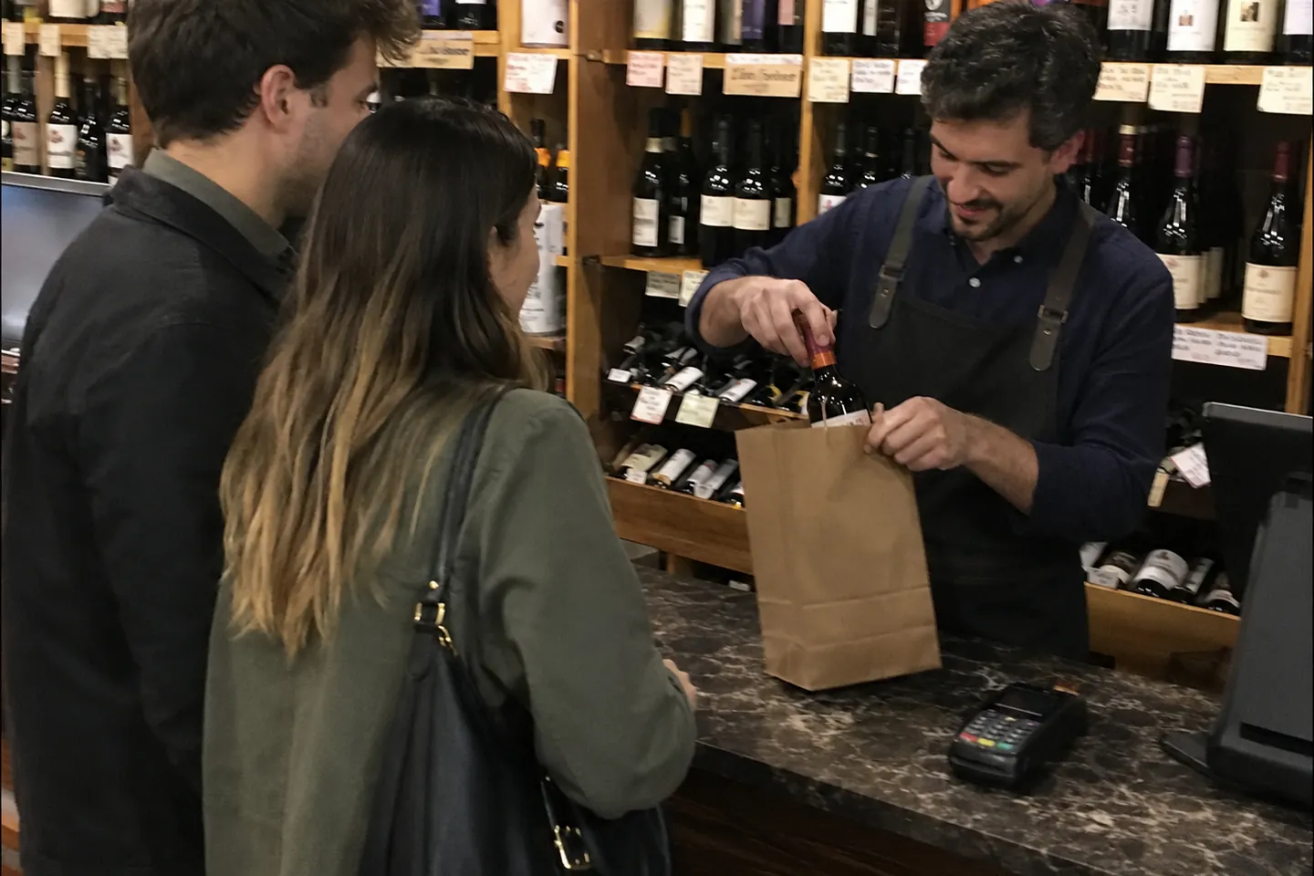 Clerk bagging bottles at the counter for two customers, with a payment terminal beside the register